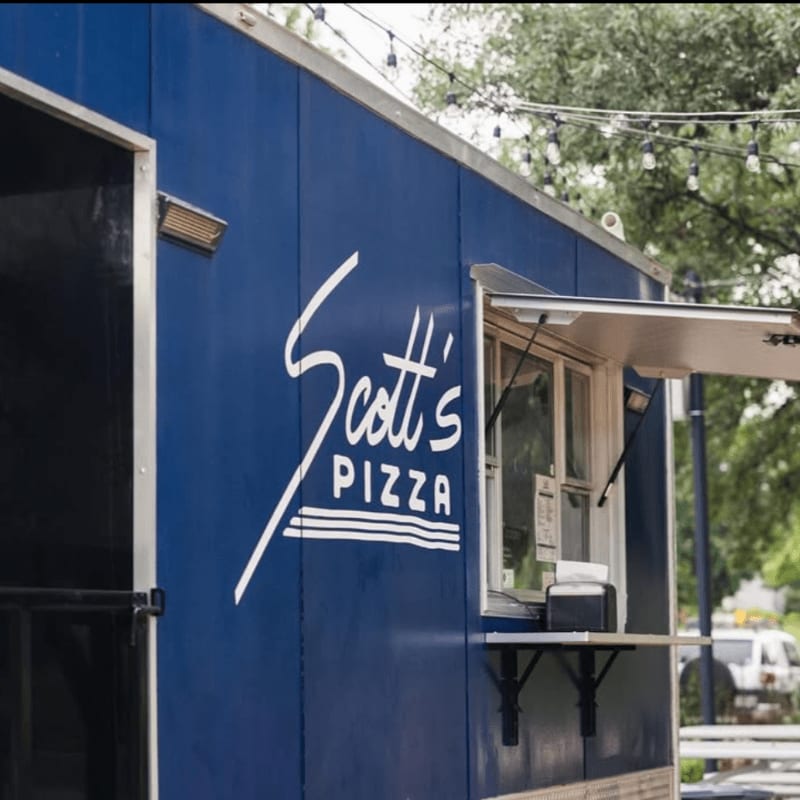 A blue Scott's Pizza food truck with an open window is parked outdoors with trees and string lights in the background.