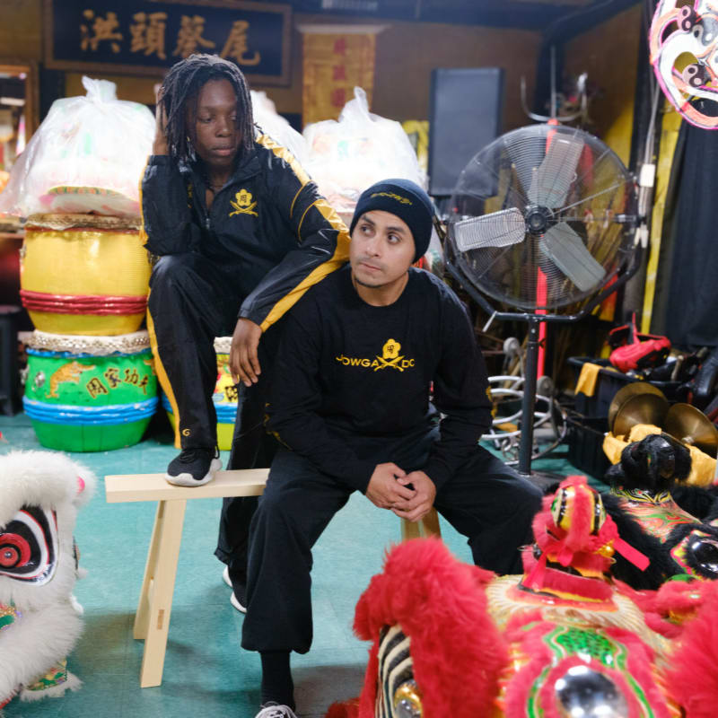 Two young men in athletic wear sit on a wooden bench in the foreground, surrounded by colorful lion dance costumes, while large drums and a fan are visible in the background.