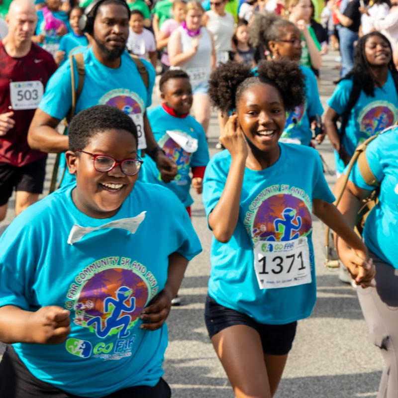 Two smiling children in blue t-shirts with race bibs run in the foreground, while a crowd of people in similar shirts participates in a race in the background.