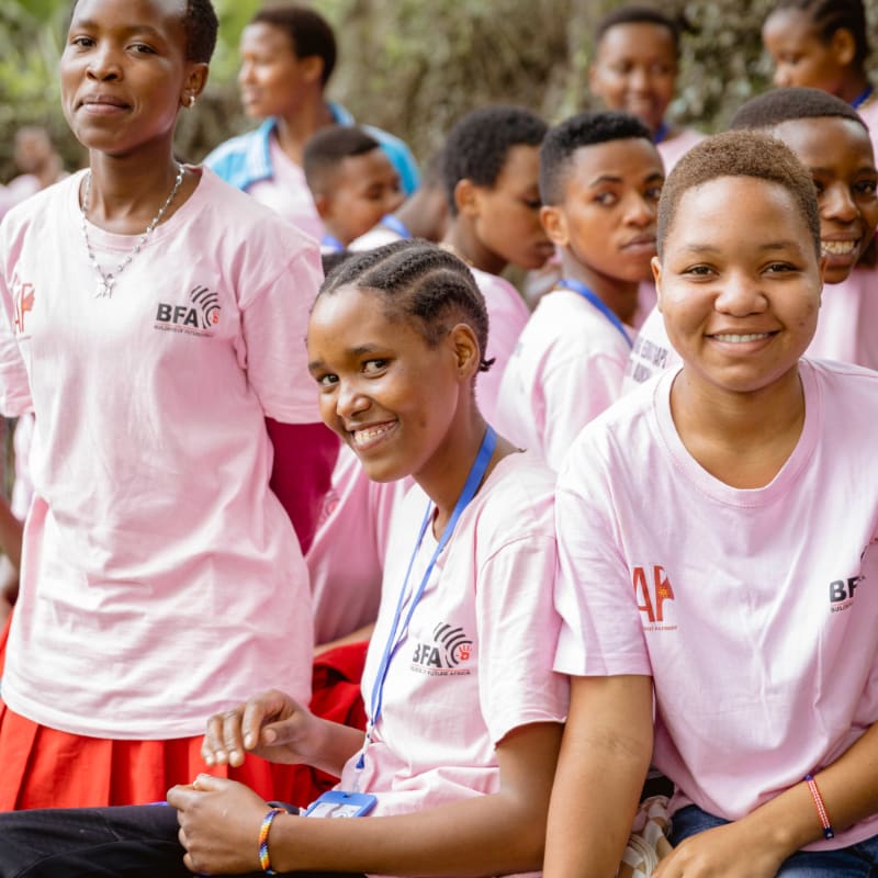 Two young women in pink t-shirts with "BFA" logos smile at the camera in the foreground, while a group of other people, also in pink shirts, are visible in the blurred background.