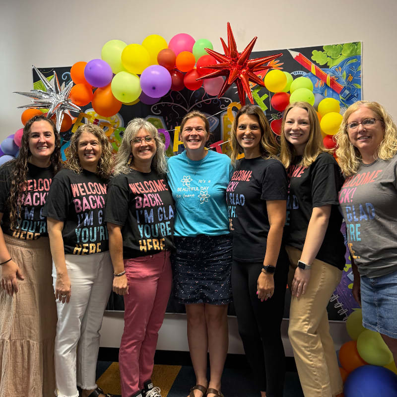 Seven women stand in front of a colorful balloon arch and a chalkboard-style mural, all wearing shirts that say "Welcome Back! I'm Glad You're Here."