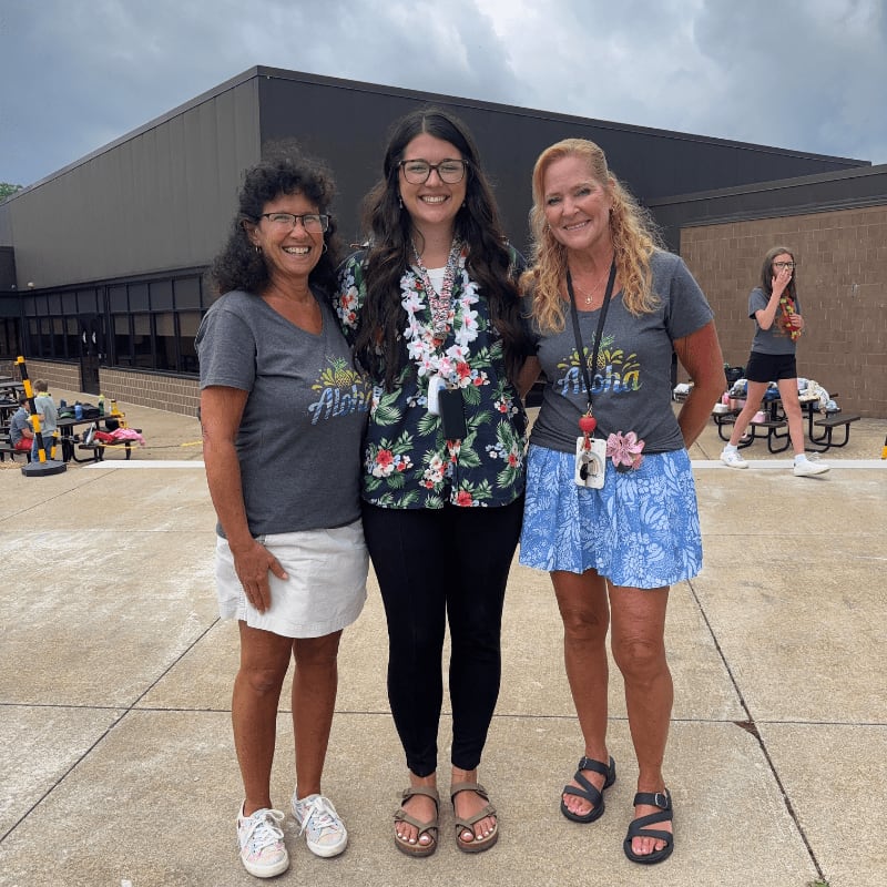 Three women in casual attire stand on a paved area in front of a building, with a blurred background showing outdoor seating and a person in the distance.