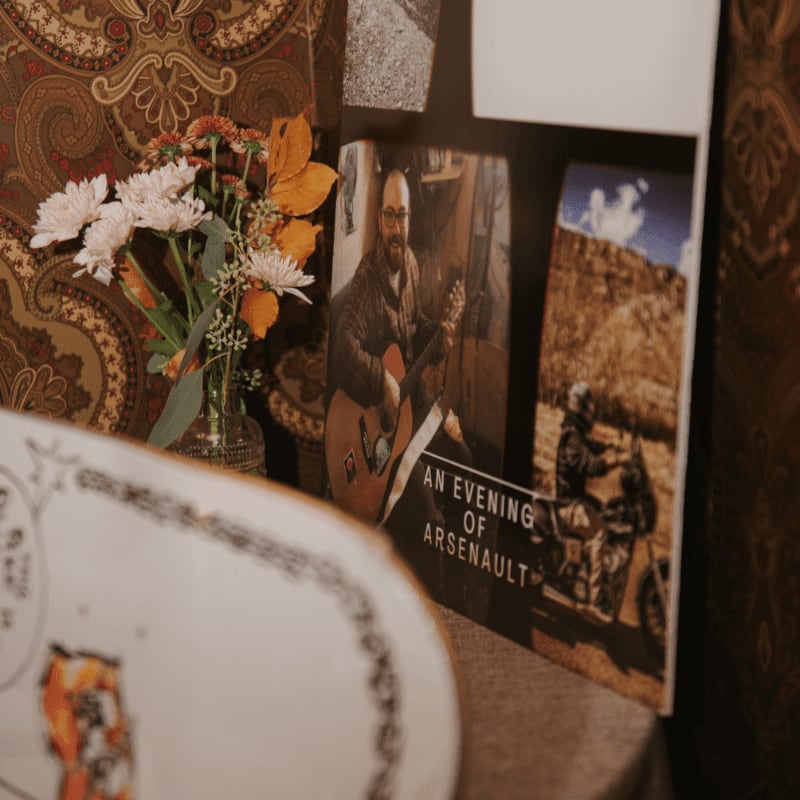 A vase of flowers sits in front of framed pictures of a man playing guitar and a motorcycle rider, all against a patterned wallpaper background.