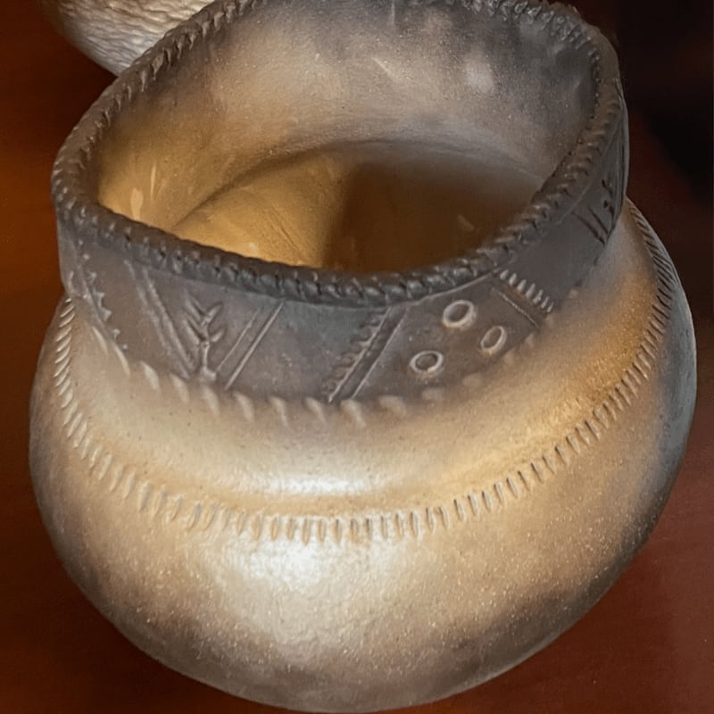 A textured, light-colored pot with decorative bands sits on a dark surface, with a blurred object in the background.