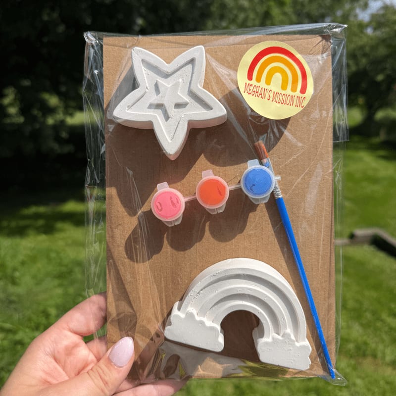 A hand holds a craft kit with a star, rainbow, and paint pots in the foreground, set against a blurred green background of trees and grass.