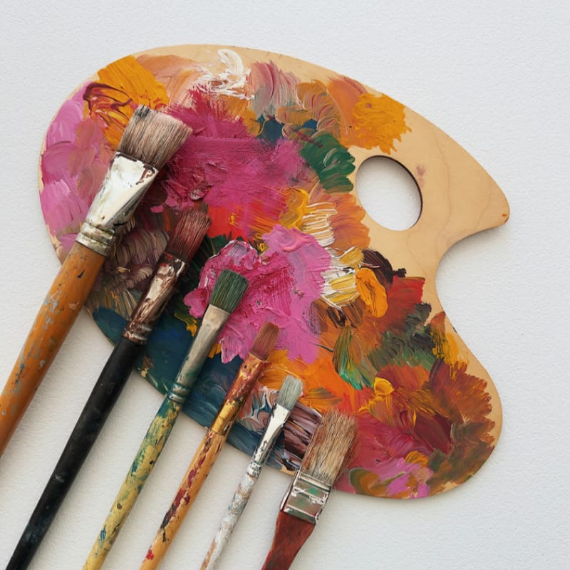 Six paintbrushes resting on a wooden palette covered in colorful oil paint