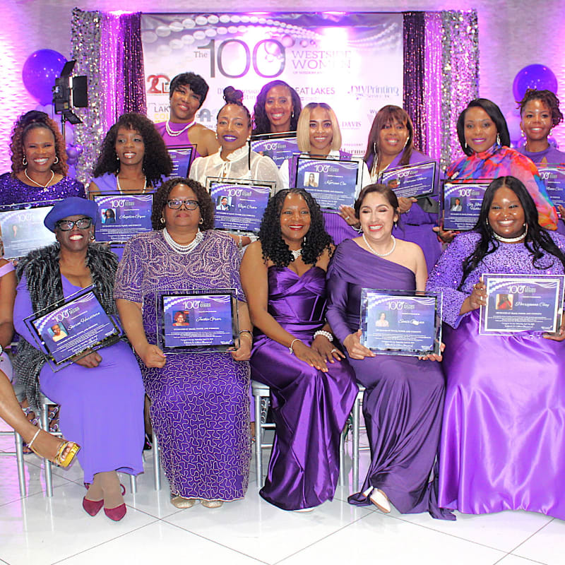 A group of women in purple dresses pose with awards in front of a sparkly backdrop.