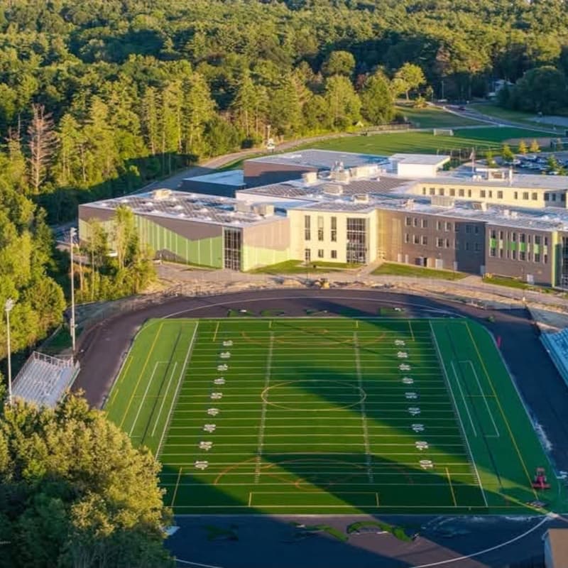 A vibrant green football field with white yard lines occupies the foreground, while a modern school building with colorful accents is nestled against a backdrop of lush green trees.