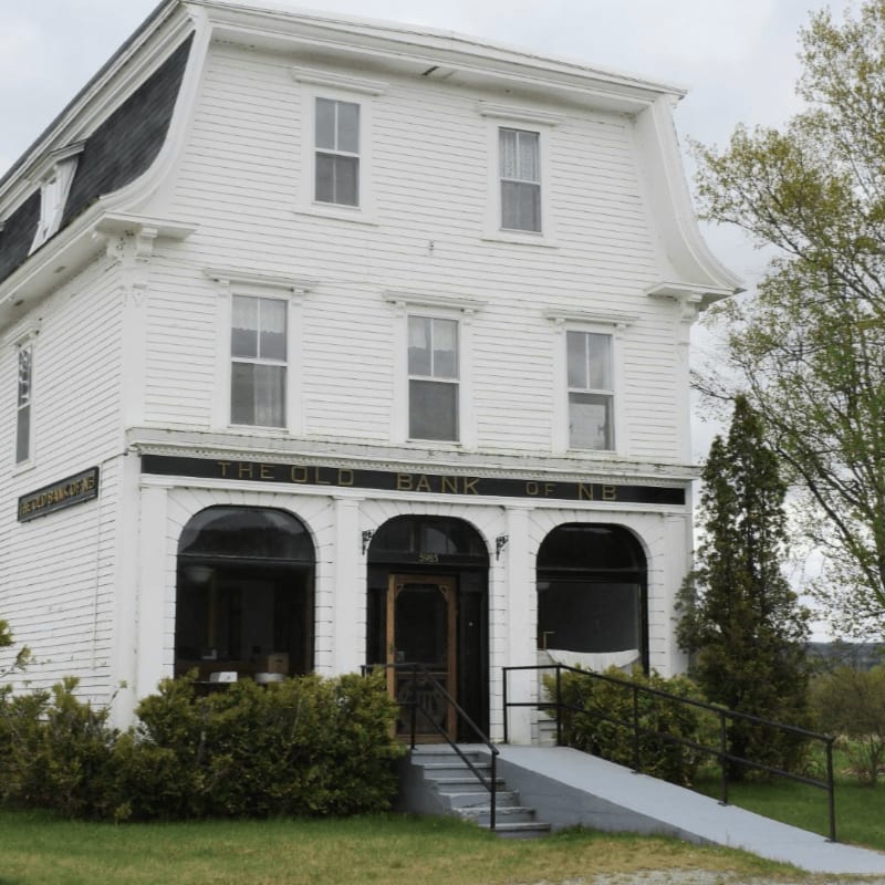 A white, historic building with arched windows and a mansard roof stands in the foreground, with lush green trees and a cloudy sky in the background.