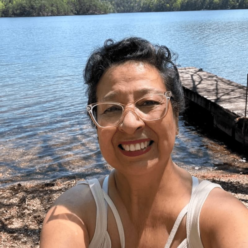 A smiling woman with glasses is in the foreground, with a wooden dock and a calm lake stretching into the background.