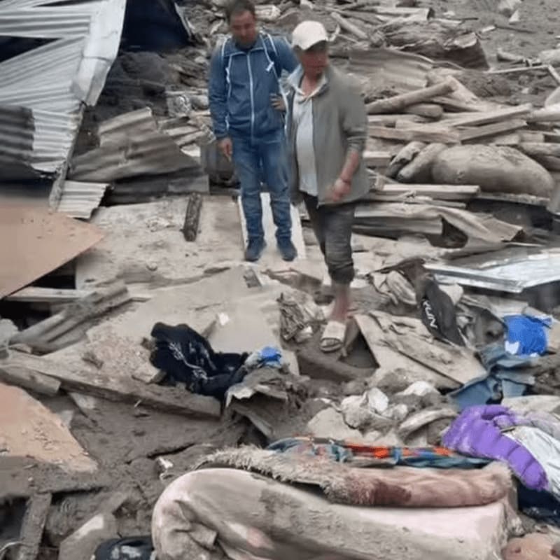 Two men stand amidst the debris of a collapsed structure, with scattered belongings and rubble filling the foreground and background.
