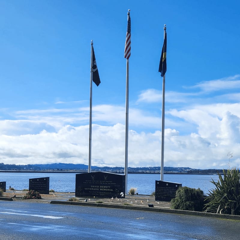 Three flags fly above a memorial with a body of water and distant hills in the background.