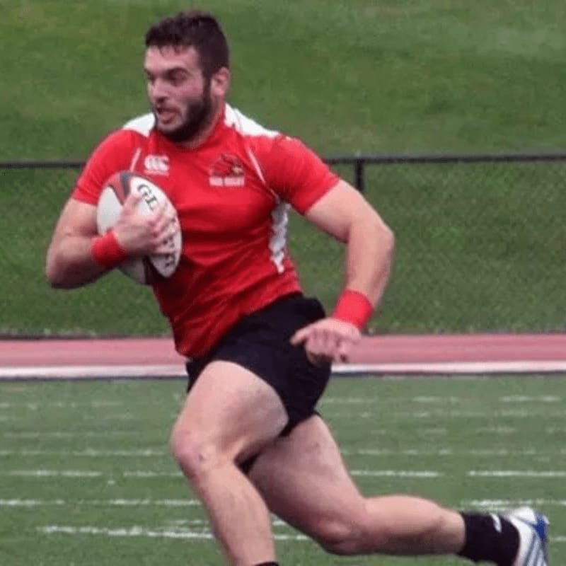 A muscular rugby player in a red jersey runs with the ball on a green field, with a blurred background of a track and fence.