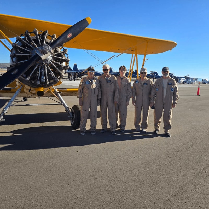 Five people in flight suits stand in front of a yellow biplane on an airfield, with other aircraft and vehicles visible in the background under a clear blue sky.