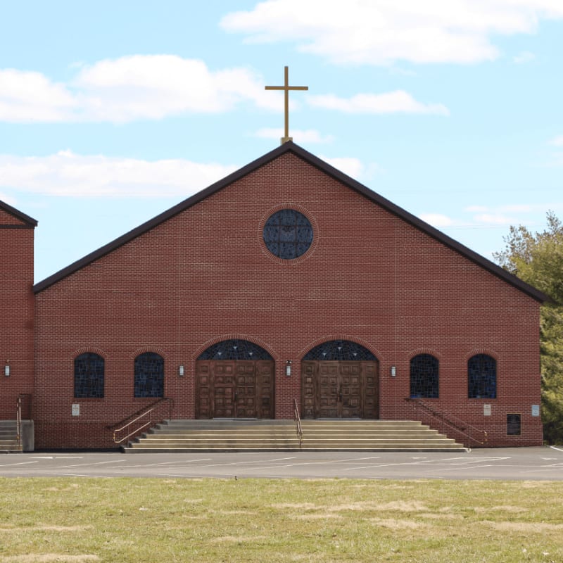 A wide, grassy lawn leads to the grand, brick facade of a church with a prominent cross atop its roof, set against a backdrop of trees and a cloudy sky.