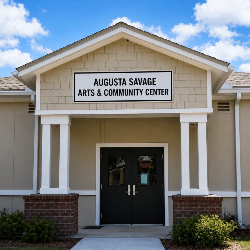 A paved walkway leads to the double doors of the Augusta Savage Arts & Community Center, a tan building with white columns, set against a bright blue sky with scattered clouds.