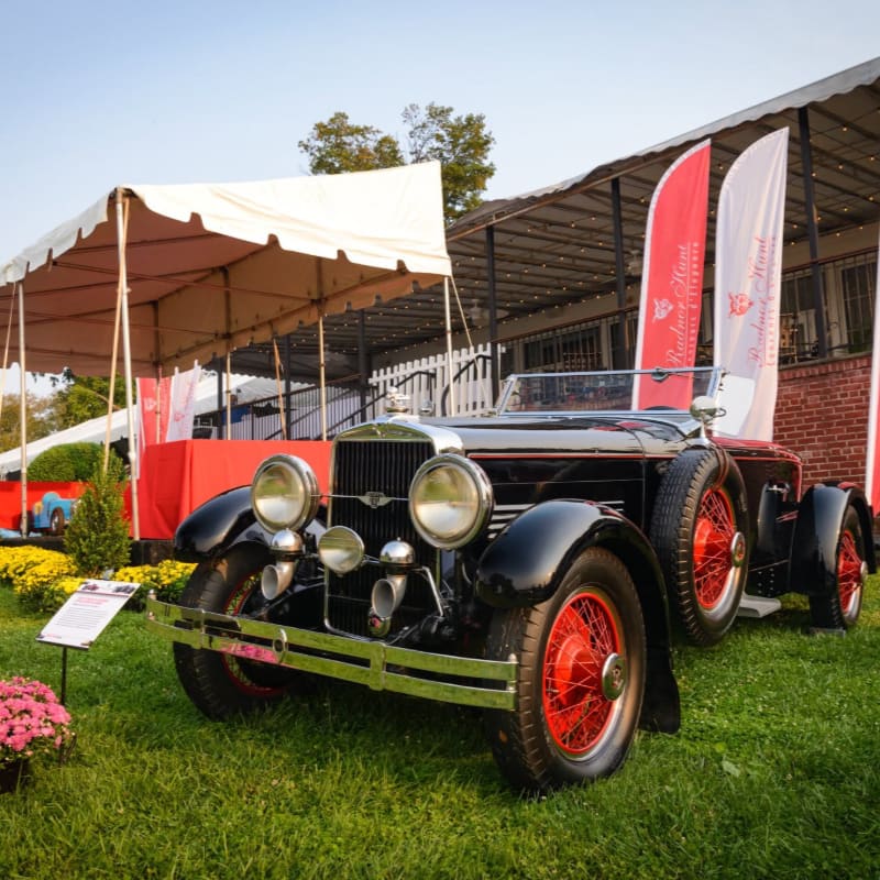 A vintage black convertible with red wheels sits on a grassy lawn in front of a white tent and a building with red and white flags.