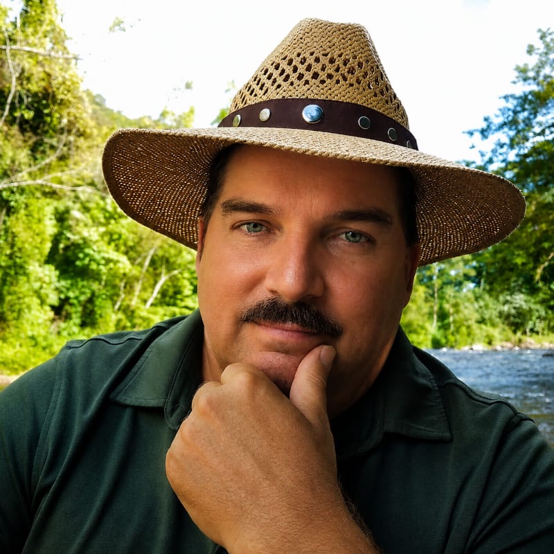 A man in a straw hat rests his chin on his hand in the foreground, with a lush, green jungle and a river in the background.