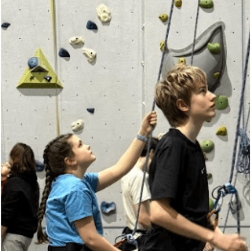 Two children, a boy and a girl, are belaying each other on a climbing wall with colorful holds and ropes in the background.