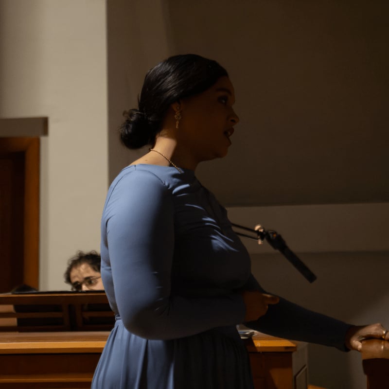 A woman in a blue dress speaks into a microphone in the foreground, with a man partially visible behind wooden pews in the background.