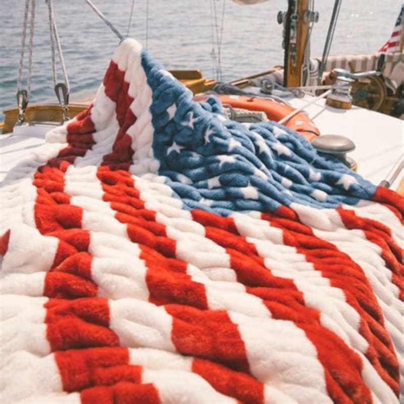 A plush American flag blanket covers the deck of a sailboat, with the ocean and sky visible in the background.