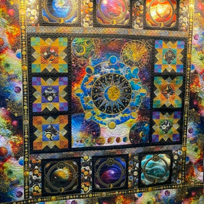 A vibrant, celestial-themed quilt with a central sun and zodiac wheel is surrounded by cosmic imagery and starry patterns.