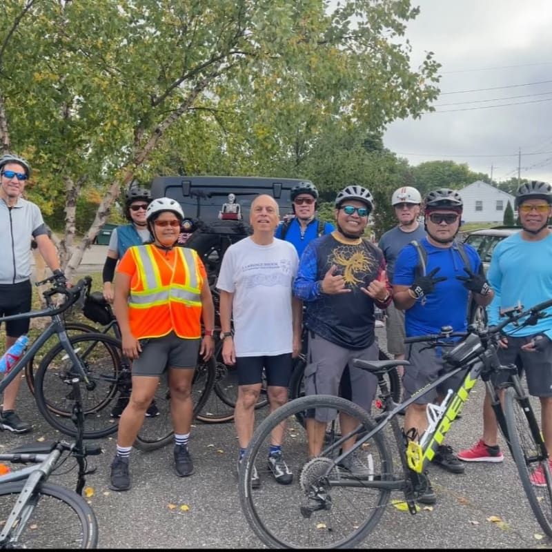 A group of cyclists in the foreground are gathered with their bikes, while a parked vehicle and houses are visible in the background.