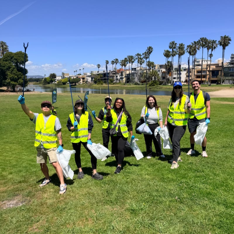 A group of volunteers in bright yellow vests stand on a grassy field holding trash bags and grabbers, with a calm body of water and palm trees lining the background.