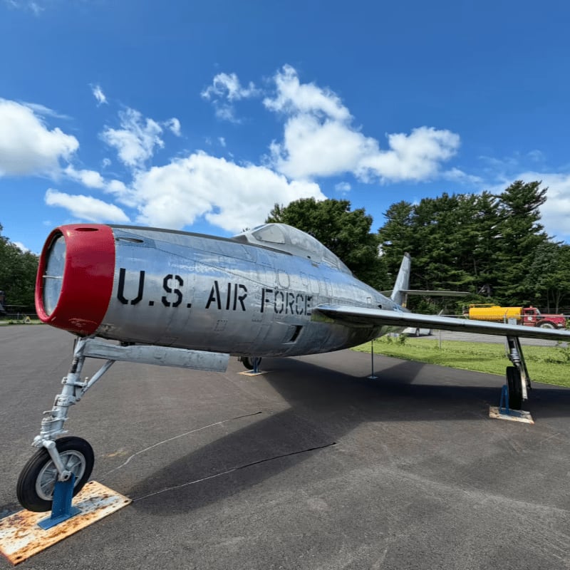 A vintage U.S. Air Force jet with a red nose sits on an asphalt pad in the foreground, with a line of trees and a bright yellow vehicle visible in the background under a cloudy blue sky.