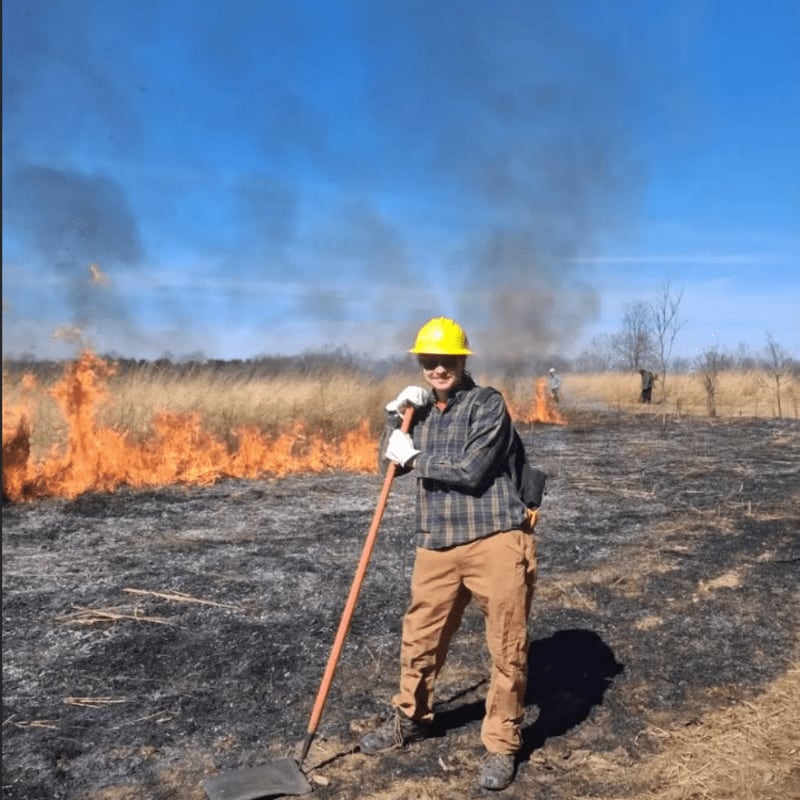 A person in a yellow hard hat and plaid shirt stands in a field of burnt grass with a rake, while a controlled fire burns in the background under a clear blue sky.