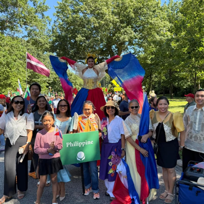 A group of people in traditional Filipino attire stand in the foreground holding a "Philippine" sign and flags, while a person in a colorful costume on stilts with large wings is elevated in the background amidst a parade with trees and other flags.