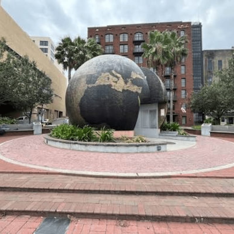 A large globe sculpture sits in a circular brick plaza with palm trees and brick buildings in the background.