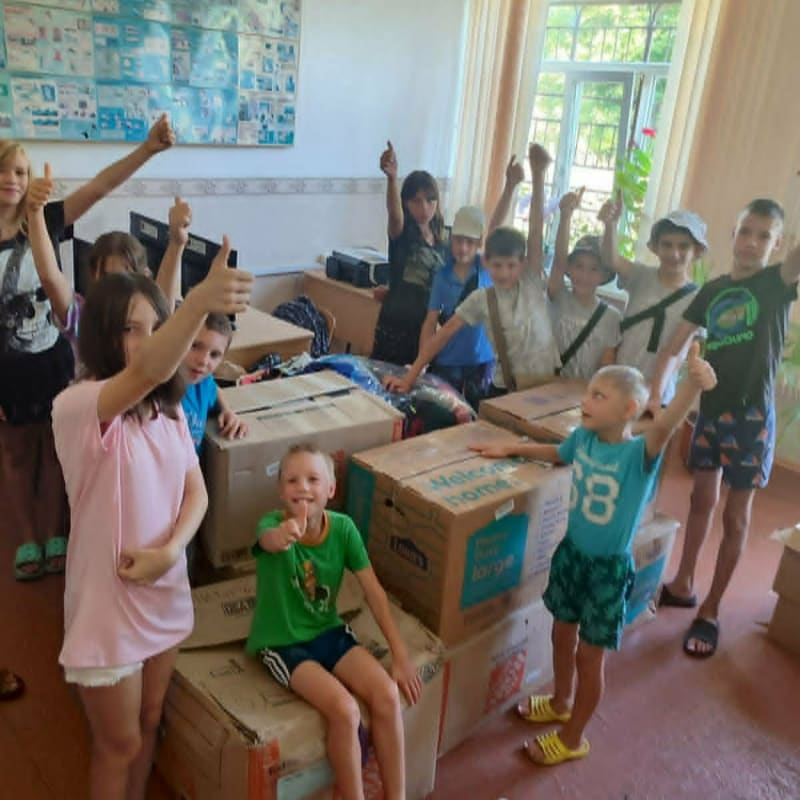 A group of children give thumbs up while posing around boxes in a brightly lit room with windows.