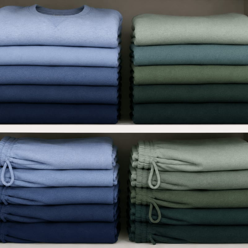 Neatly folded stacks of blue and green sweatshirts and sweatpants are organized on two shelves against a plain background.