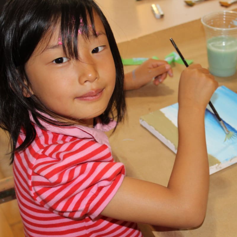 A young girl in a pink striped shirt paints a canvas with a brush in her right hand, while art supplies and a cup of green liquid are visible in the background.