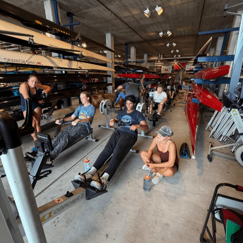 In the foreground, several people are using rowing machines, while the background reveals racks filled with rowing shells.
