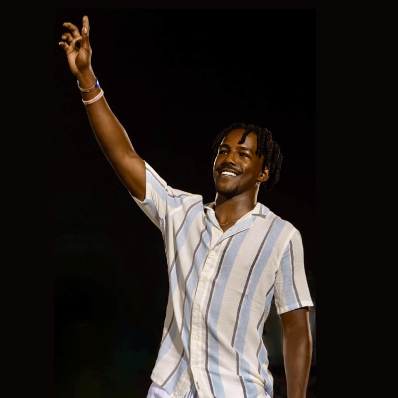 A young man in a striped shirt raises his hand against a dark background.