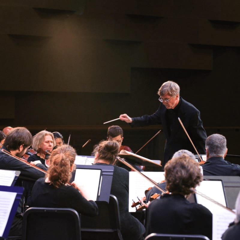 A conductor leads an orchestra in a concert hall, with musicians playing their instruments in the foreground and the stage lighting illuminating the background.