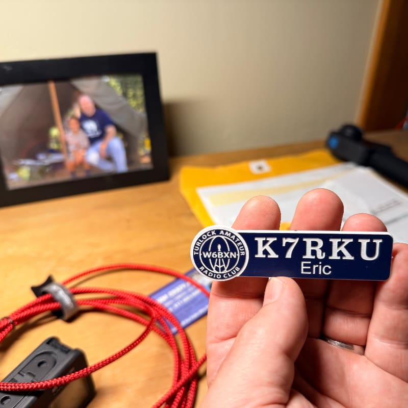A hand holds a name tag with "K7RKU Eric" on it, with a framed photo of people in a tent and red wires in the foreground, and a yellow envelope and a black object in the background.
