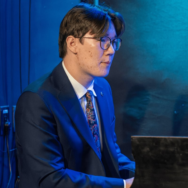 A man in a suit and tie plays a piano with a blue background.