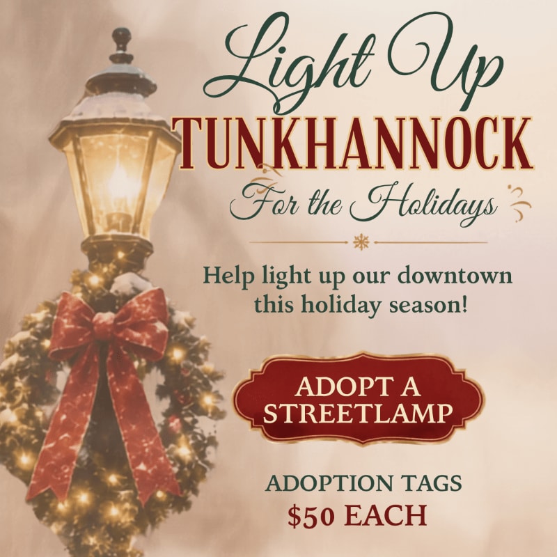 A festive streetlamp adorned with a wreath and bow stands in the foreground against a softly lit, textured background promoting the "Light Up Tunkhannock For the Holidays" event.