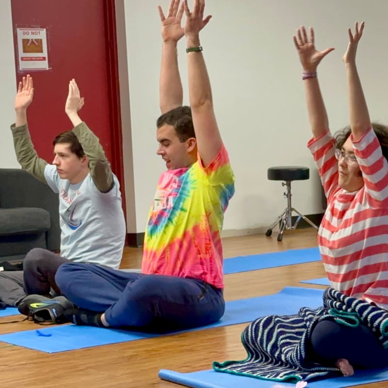 Three people are meditating on yoga mats in the foreground, with a red wall and a stool visible in the background.