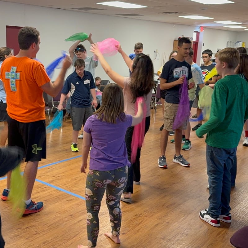 A group of young people in the foreground are joyfully dancing with colorful scarves, while others in the background watch and participate in a brightly lit room.
