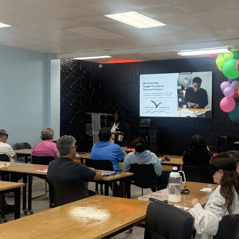 In the foreground, people sit at desks facing a presenter, while the background features a large screen displaying a presentation on personal finance and colorful balloons.