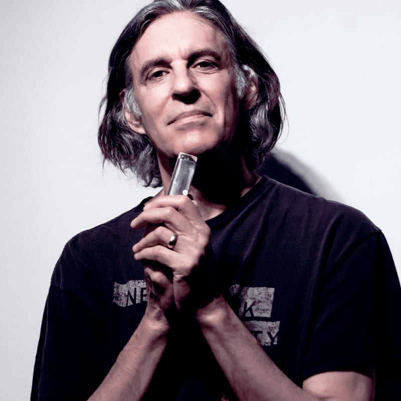 A man with long, graying hair holds a harmonica to his lips in front of a plain, white background.