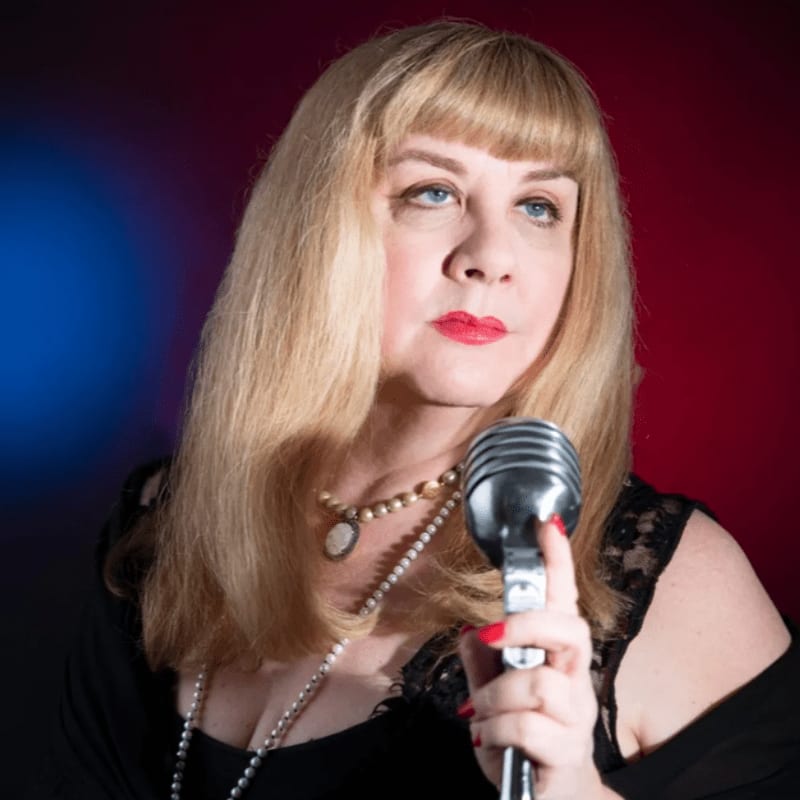 A woman with blonde hair and pearls holds a vintage microphone in front of a red and blue spotlighted background.