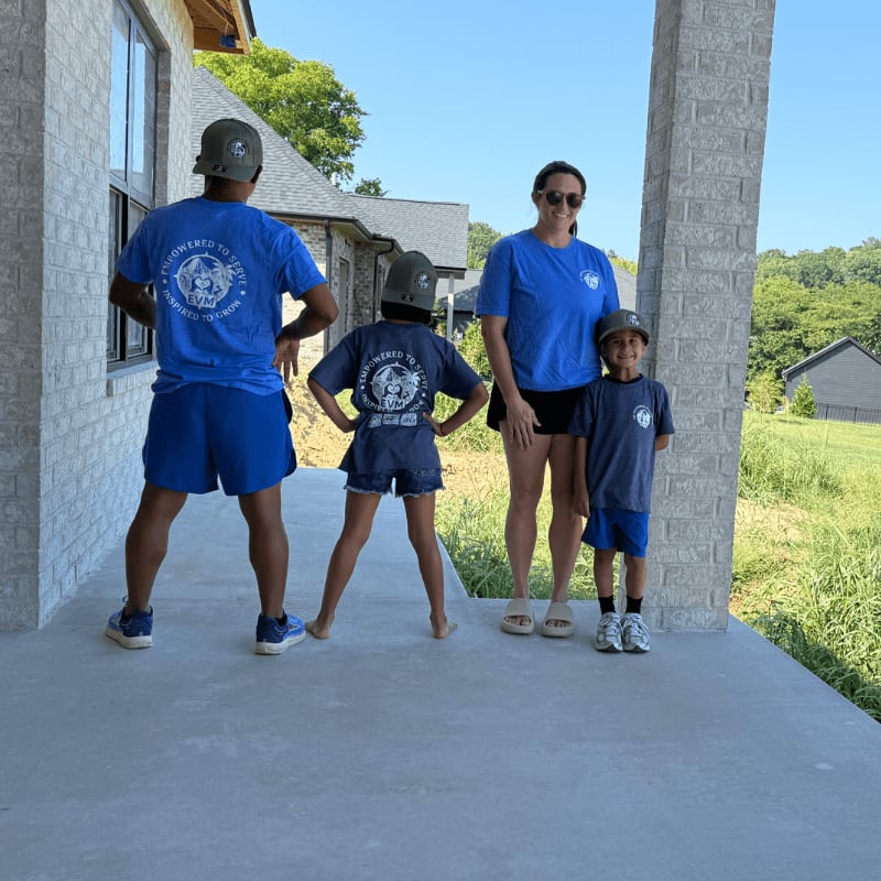 A family of four, dressed in matching blue shirts and shorts, poses on a concrete porch in front of a house under construction, with a lush green landscape in the background.