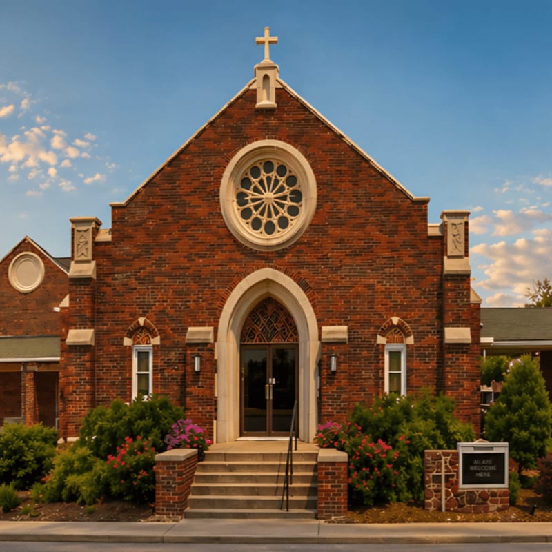 A brick church with a rose window and cross stands before a blue sky with fluffy clouds, surrounded by lush green bushes and a stone sign.