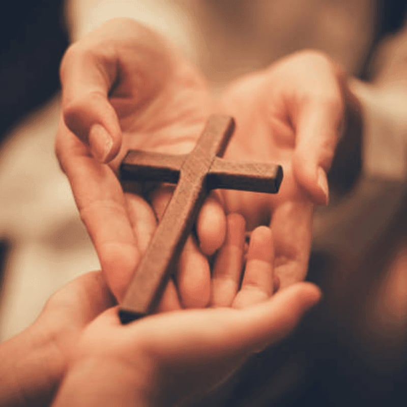 Two pairs of hands gently hold a wooden cross in the foreground, with a soft, blurred background.