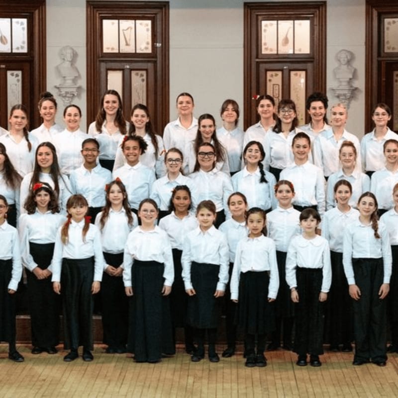 A large choir of children and adults in white shirts and black bottoms stand in rows in front of a wall with ornate doors and busts.